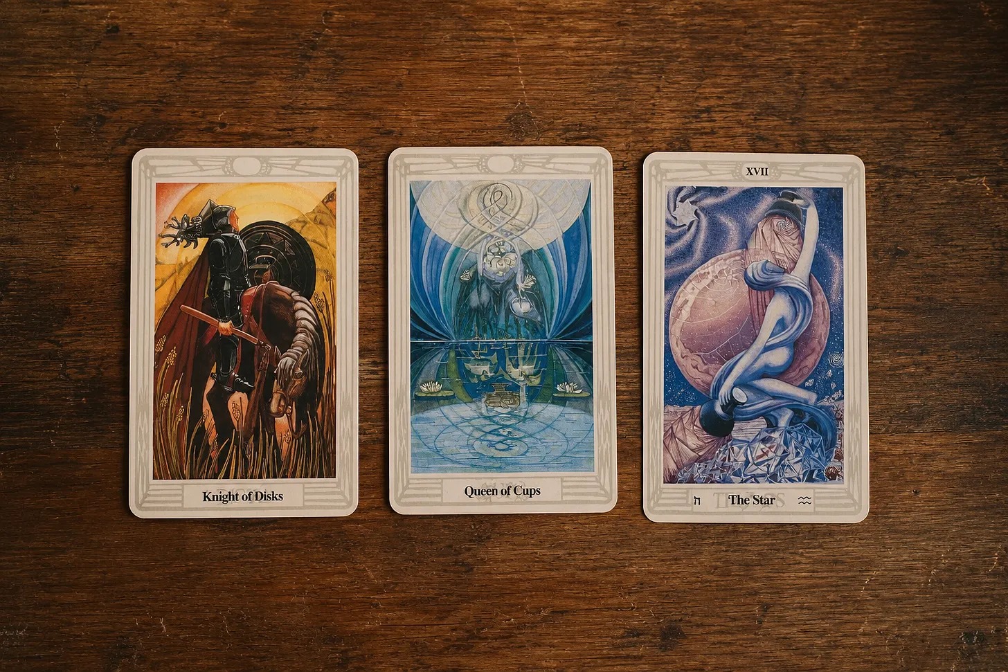 Three cards from M's own Thoth deck on a wooden table: the Knight of Disks, the Queen of Cups and The Star.